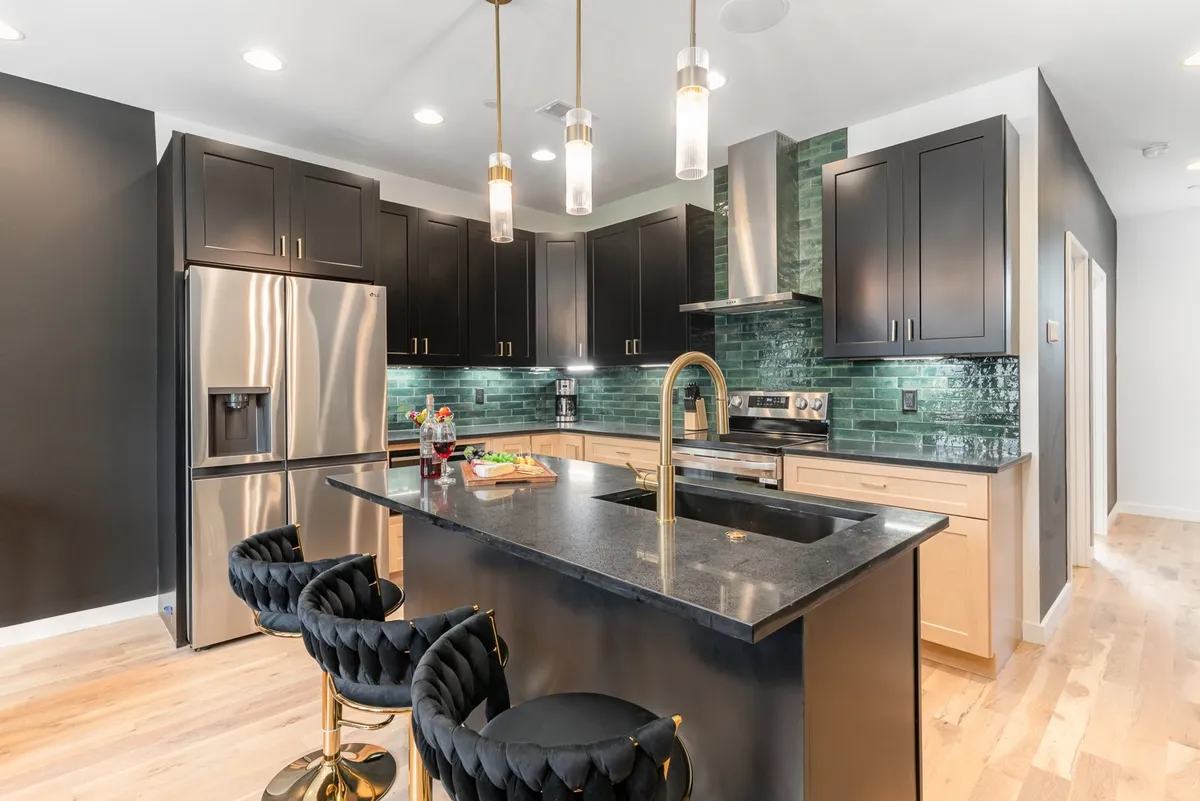 A sophisticated kitchen with black cabinetry, a green tile backsplash, a central island with seating, and gold fixtures. The setup is both chef-ready and visually striking, inviting guests to gather and snack or mingle while cooking.