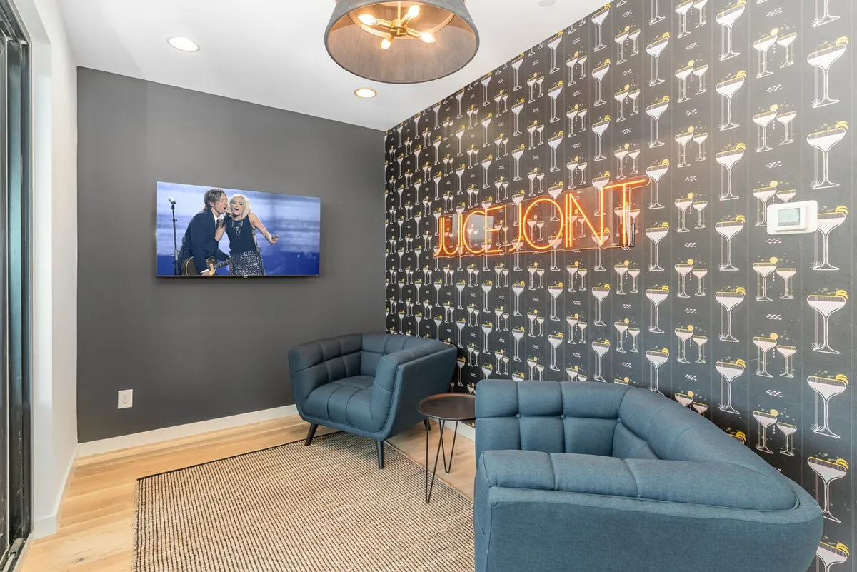 A lively lounge space featuring a neon sign reading 'JUICE JOINT,' which immediately evokes the Prohibition-era speakeasy vibe. Comfortable blue armchairs and a television displaying a performance sit against a stylish patterned wallpaper. The room is arranged for socializing, watching movies, or relaxing with a drink.