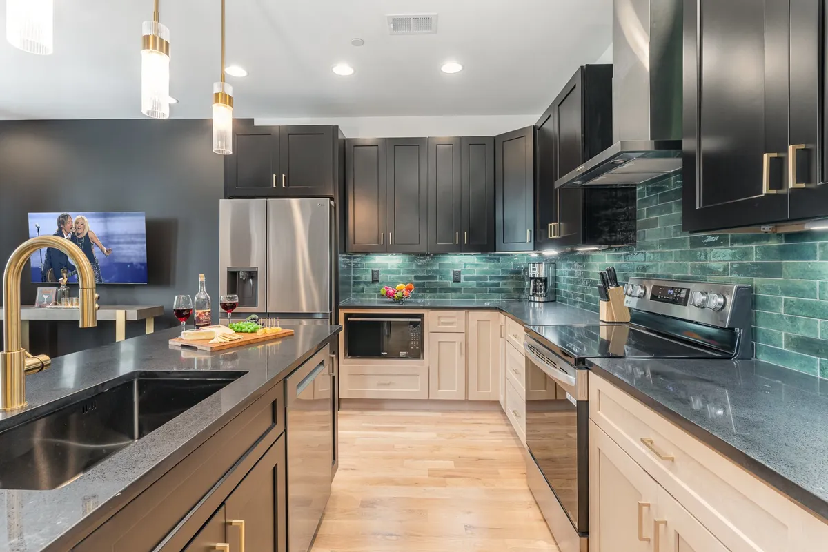 A modern kitchen featuring a central island, black cabinets, a vibrant green backsplash, and stainless steel appliances. Decorative items add personality to the open counters, and the setup radiates a welcoming, social atmosphere ideal for cooking and entertaining.