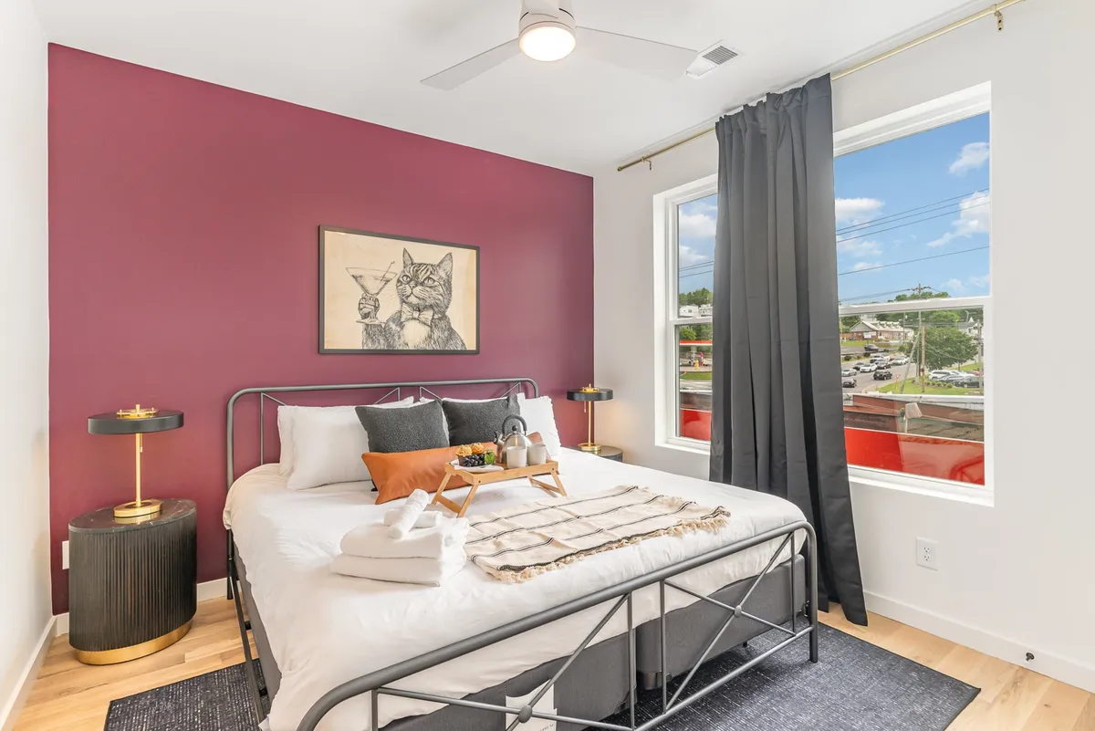 A bedroom scene with a decorative bed, whimsical cat artwork, large windows offering a pleasant view, and a cozy nightstand with a lamp. The room is suffused with natural light and warmth, blending style and comfort.