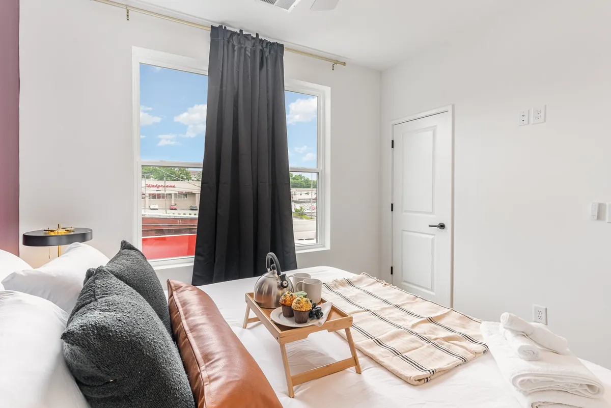 A serene bedroom scene with a bed layered in decorative pillows, a breakfast tray with a teapot and snacks, and large windows streaming with natural light. The mood is tranquil, ideal for relaxation and quiet mornings.