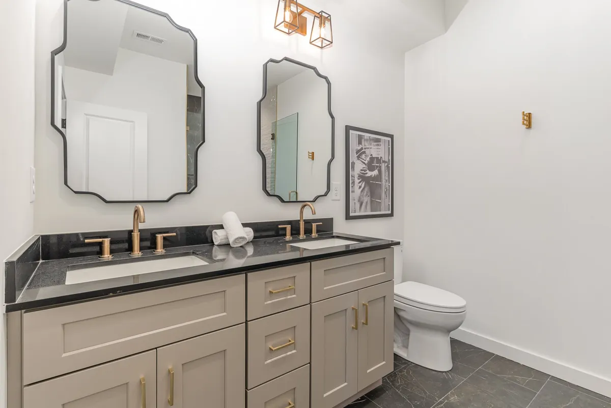 An elegant bathroom space featuring dual mirrors, a black granite countertop with gold fixtures, and framed artwork. The setting is refined and refreshing, perfect for morning routines or self-care.