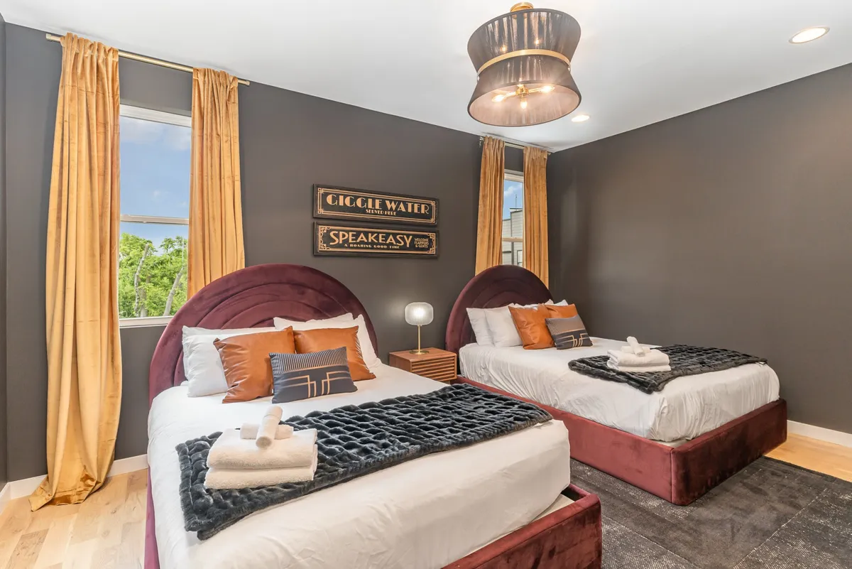 This image features two beds with plush headboards and decorative throw pillows, enhanced by a hanging light fixture and vintage-themed wall art. The overall palette and decor feel both cozy and stylish, suggesting a blend of comfort and designer aesthetics.