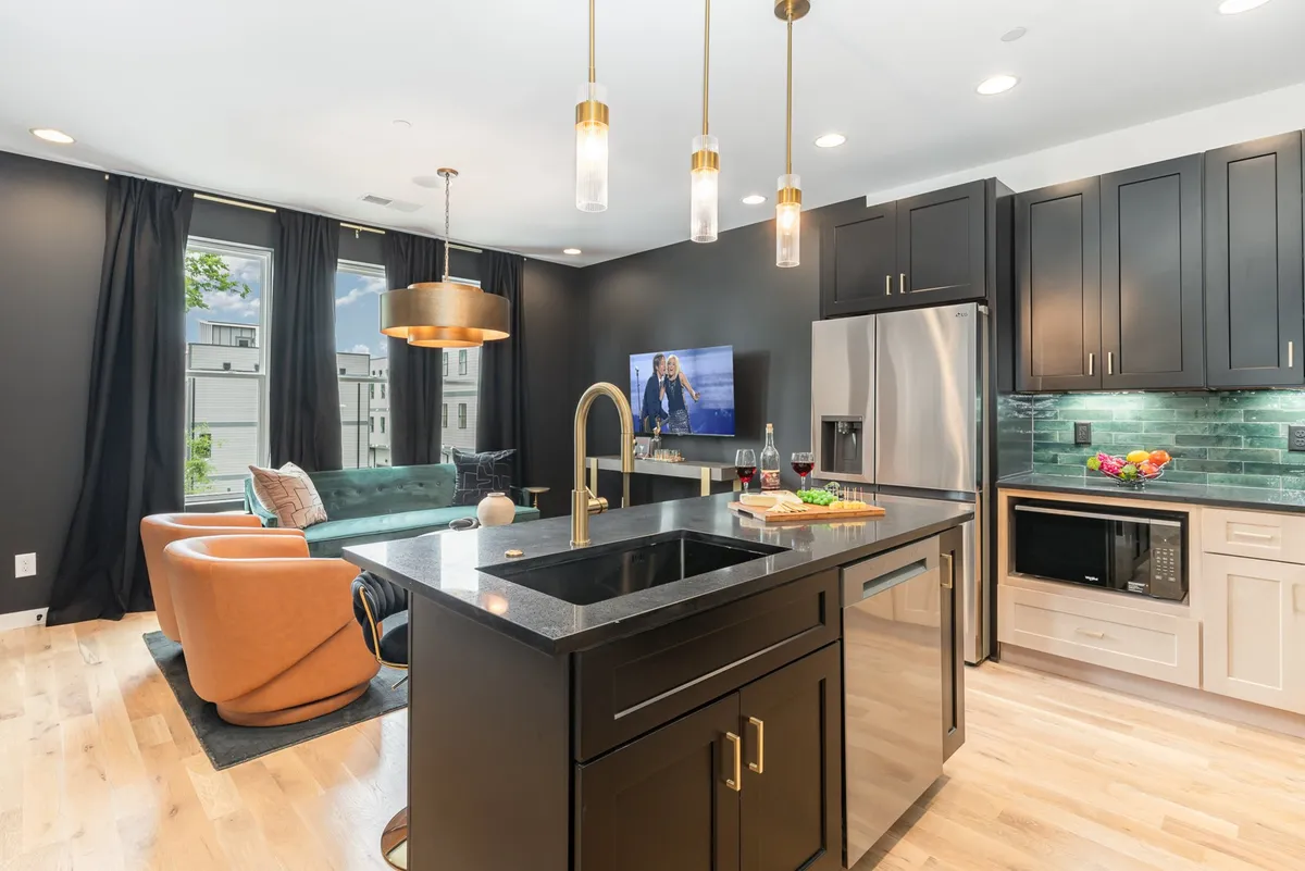 An elegant interior view with a kitchen island, stylish black cabinetry, gold pendant lights, and a comfortable seating area adjacent to large windows. The arrangement encourages interaction between the kitchen and lounge areas, underscoring a seamless flow between cooking and entertaining.