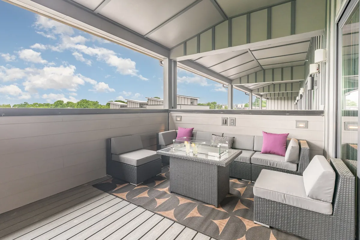 This image displays a comfortable outdoor seating area on a balcony or deck, featuring a glass coffee table and plush decorative pillows. The space is bathed in warm, natural light and suggests an inviting spot for relaxation, entertaining, or enjoying drinks at sunset. The view hints at the transition from indoors to outdoors, emphasizing the private aspect of the deck.