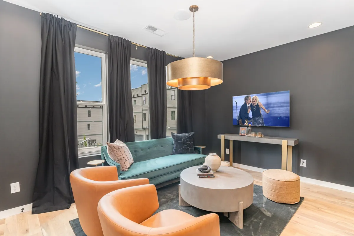 A modern main living room featuring a turquoise sofa, round coffee table, large windows allowing natural light, and a wall-mounted TV. The open layout and inviting seating make this space ideal for group gatherings, watching TV, or relaxing together.